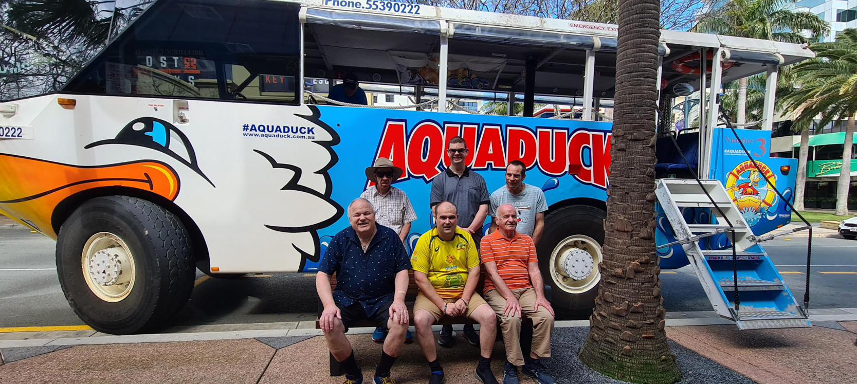 Six male clients (3 seated, 3 standing) in front of the Aqua Duck - a white and blue tour bus that also turns into a boat. 