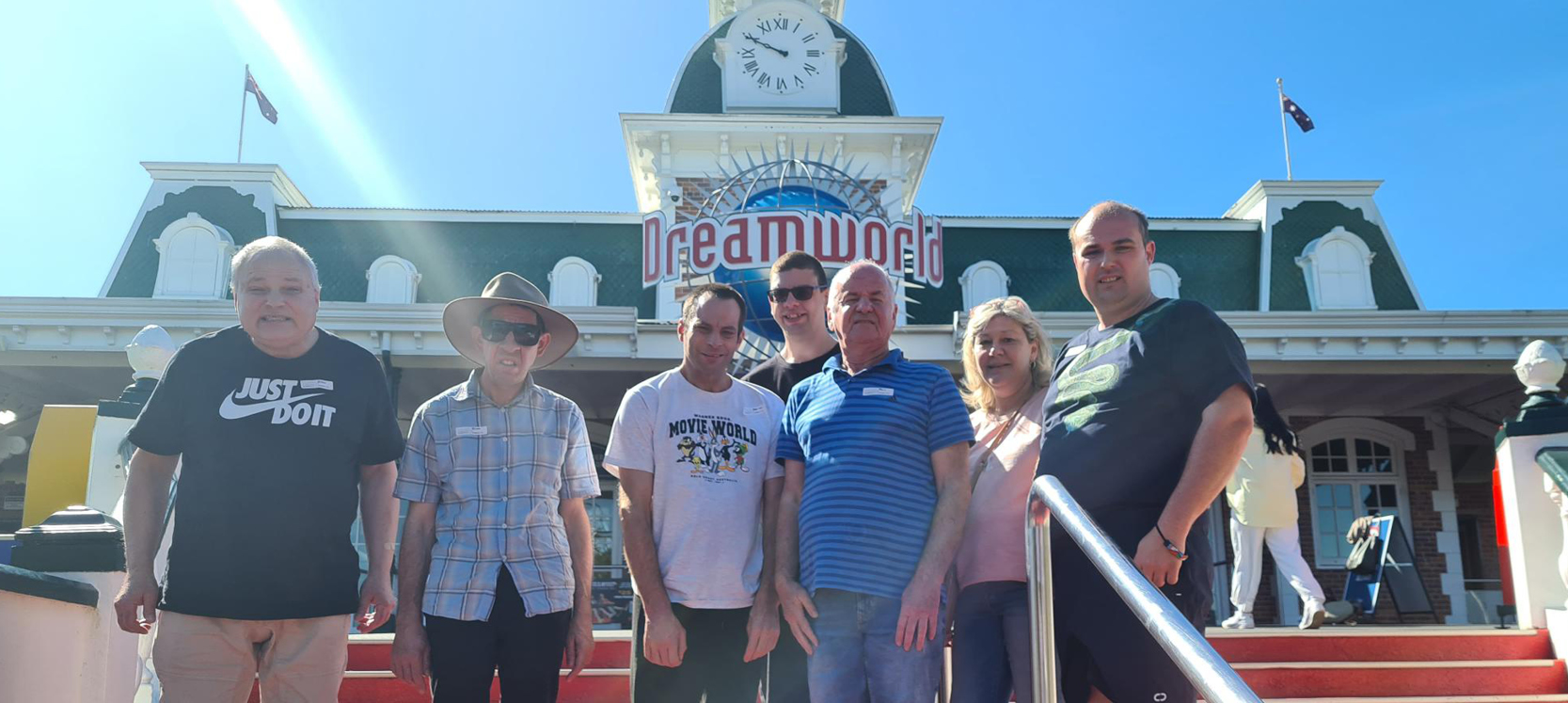 Six male clients and one female Orana staff member smiling and standing in front of the DreamWorld sign in Queensland. 