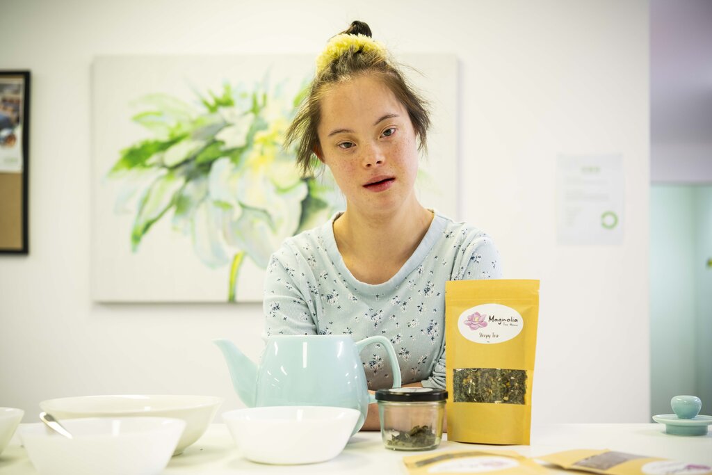 A woman in a blue top poses with a teapot and loose leaf tea in a bag. 