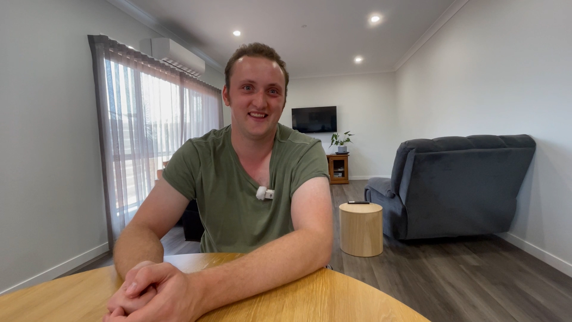 Man in green t-shirt sits at a wooden table smiling for the camera