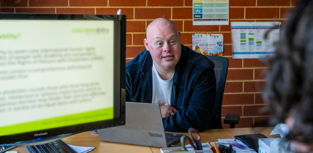 Man in white t-shirt with black jacket sits at a desk with a computer