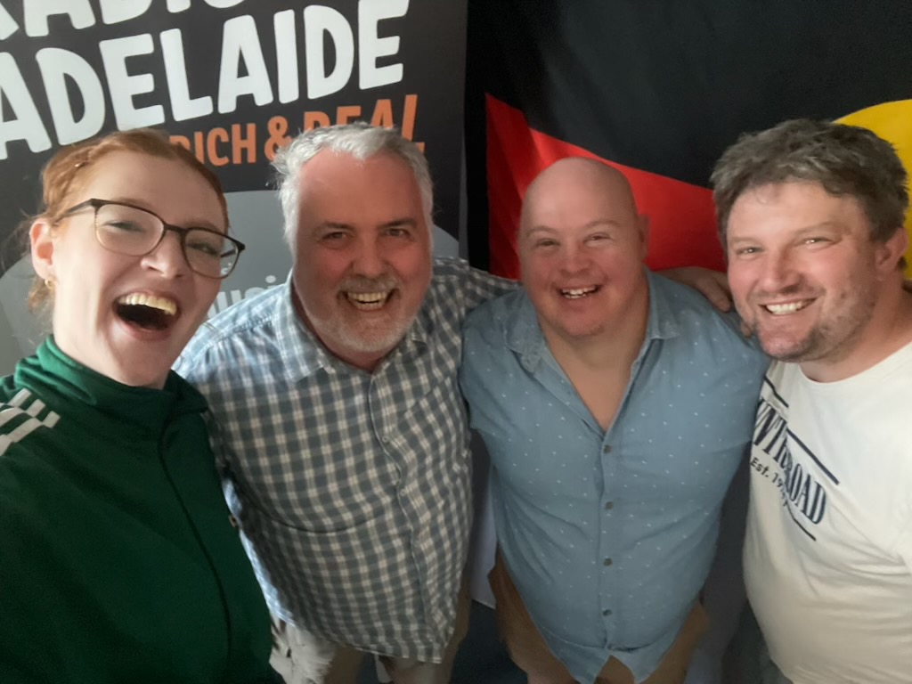 Four people smile for the camera in front of a Radio Adelaide sign
