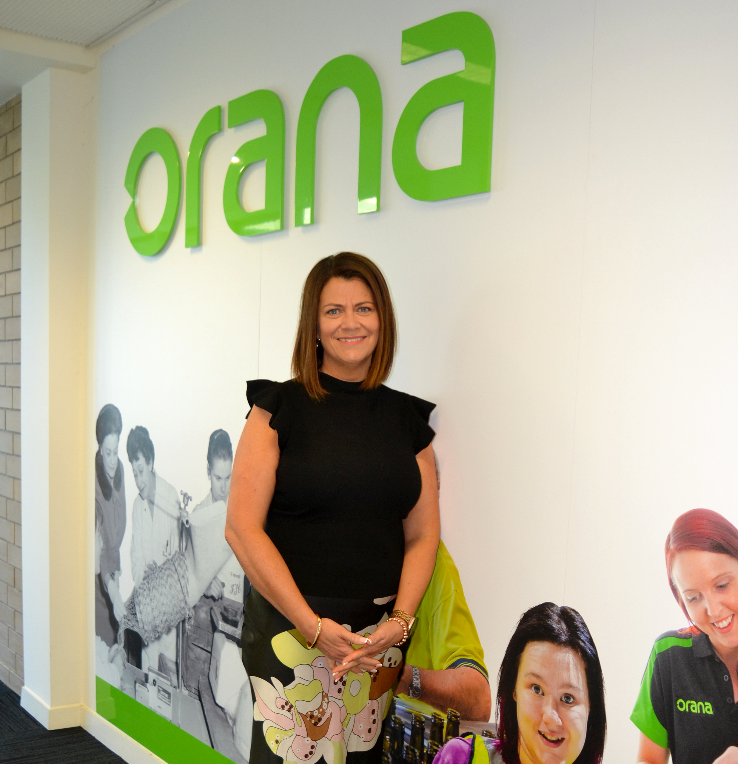 Deb Dutton in a black top and long shirt in front of a green Orana sign