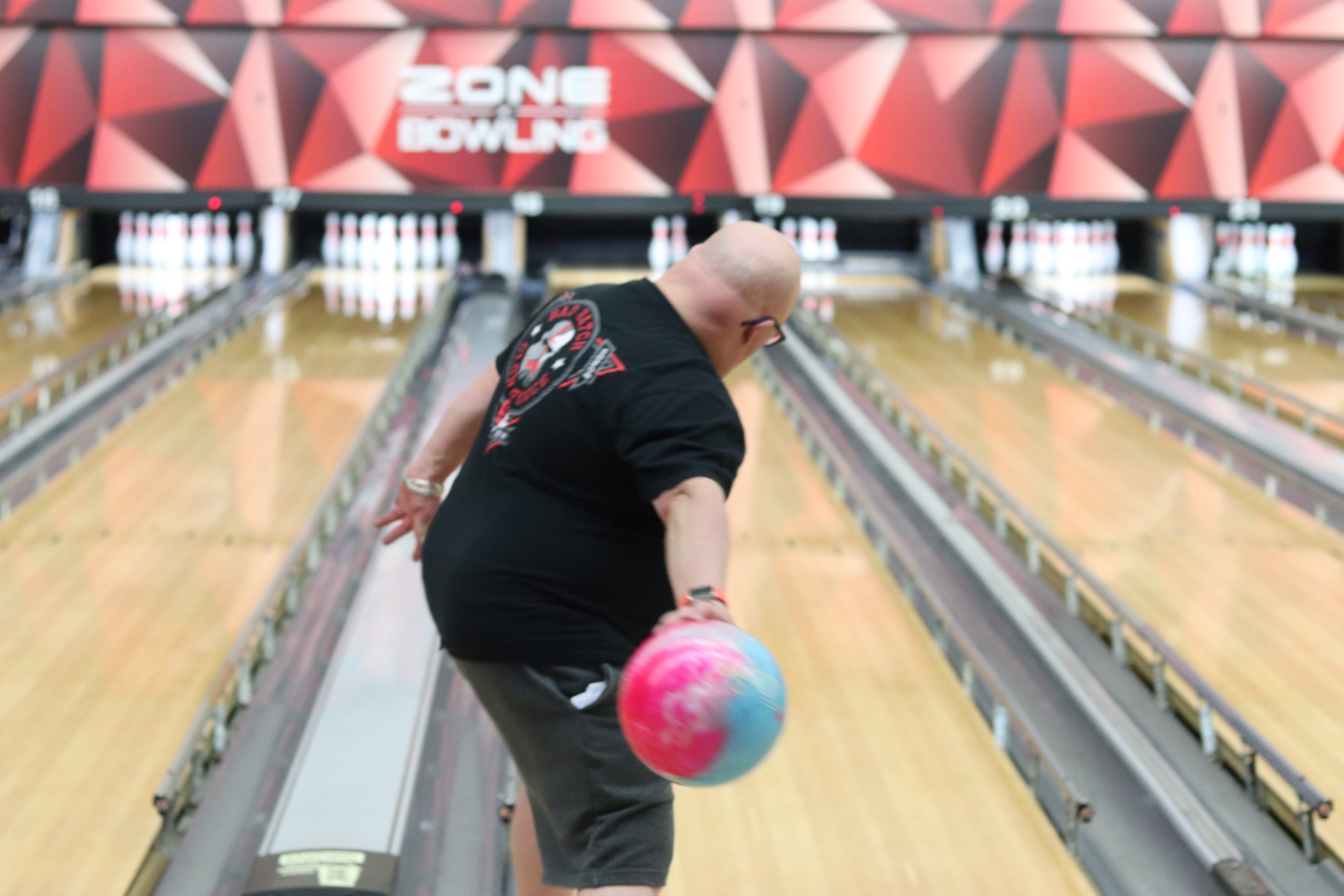 Man in black shirt with pink and blue bowling ball about to bowl down a bowling alley