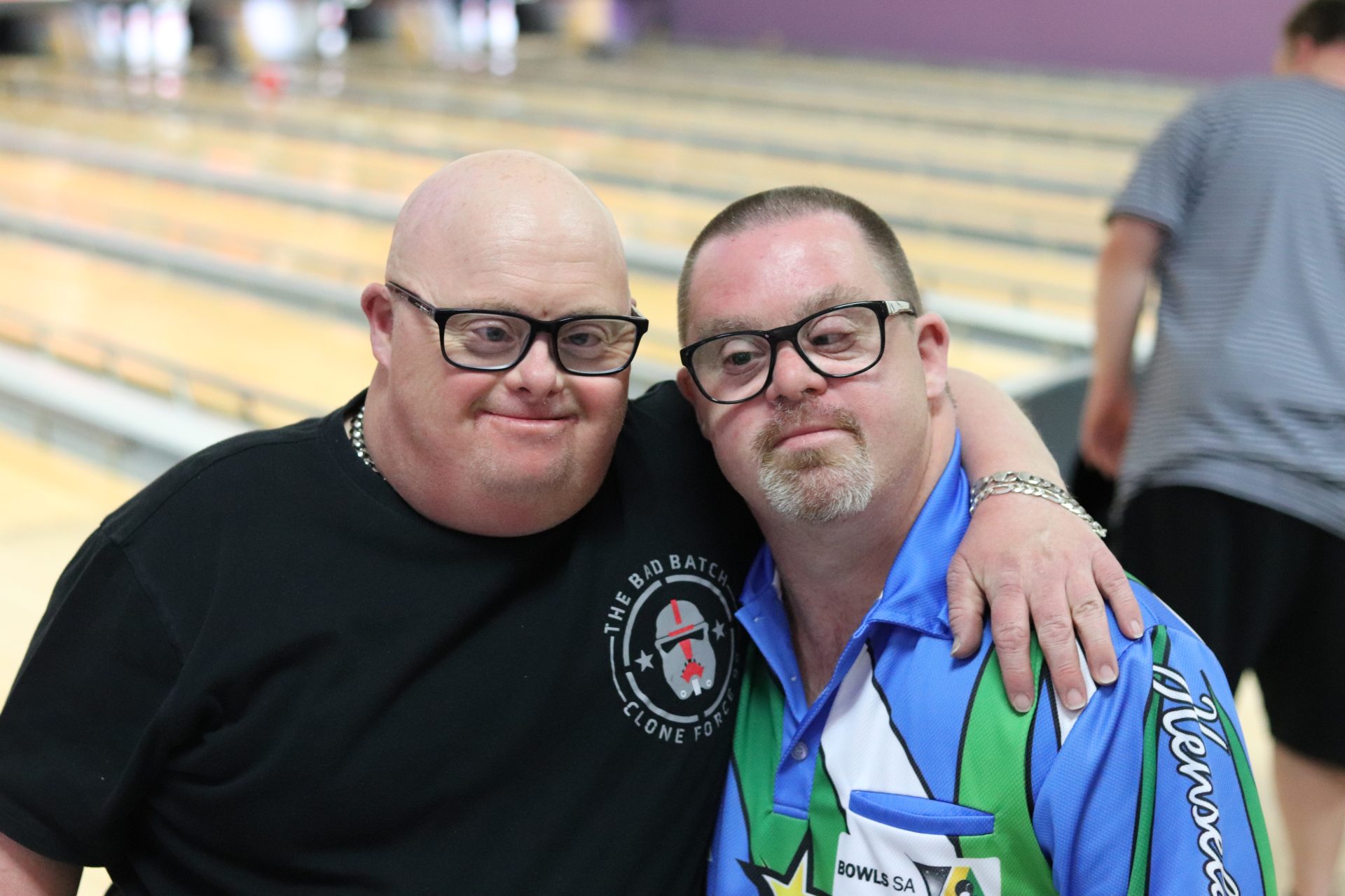 Two men, one in a black t-shirt and one in a blue, green and white shirt pose for the camera