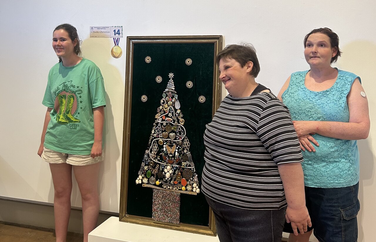 Three women stand in front of a Christmas tree made out of various craft pieces