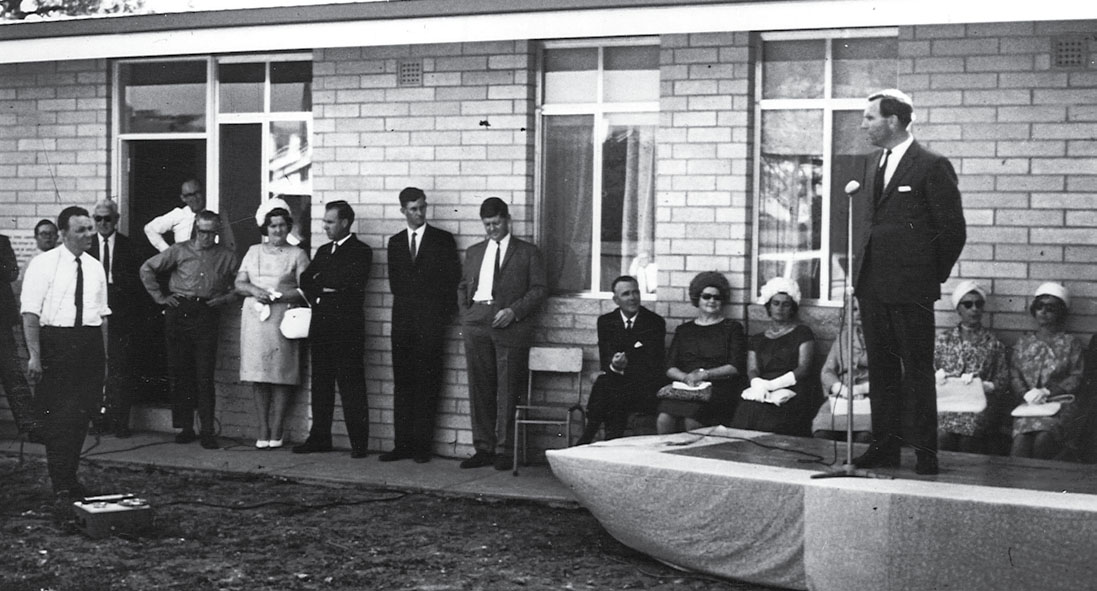A black and white photo of a man in suit making an announcement to a large group of people in front of a house